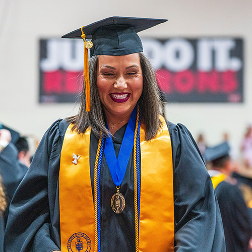 female student with several pieces of regalia and medals, smiling while walking at a graduation ceremony