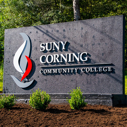 the SUNY corning community college welcome sign on a sunny day. The welcome sign is black with white letters and the CCC logo is in white, gray and red. Small green shrubs are underneath the sign and in brown mulch.