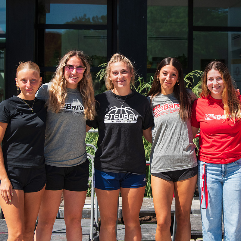 A group of five people stand closely together outdoors in bright sunlight. They are posing in a line with arms around one another. All are wearing casual athletic clothing, including T‑shirts with printed text such as “Baron,” “Steuben Basketball,” and shorts or jeans. Behind them is a building with large windows and greenery visible in planters.