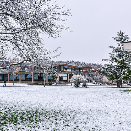 A modern campus building surrounded by snow-covered trees and grass on a winter day. A person in a blue coat walks along a path in the distance. The sky is overcast, and the scene shows a mix of green grass peeking through the snow.